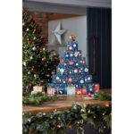Adventskalender-Turm auf Holztisch neben Weihnachtsbaum und Christbaumkugeln