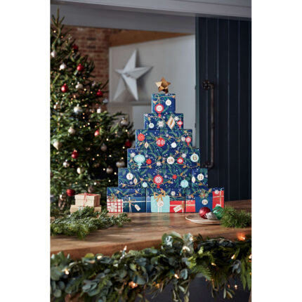 Adventskalender-Turm auf Holztisch neben Weihnachtsbaum und Christbaumkugeln