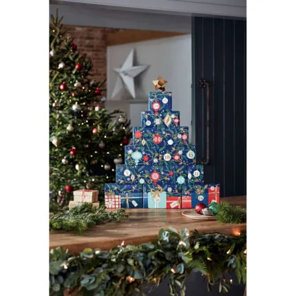 Adventskalender-Turm auf Holztisch neben Weihnachtsbaum und Christbaumkugeln