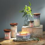 WoodWick Trilogy Kerze neben weiteren brennenden Kerzen und Pflanze