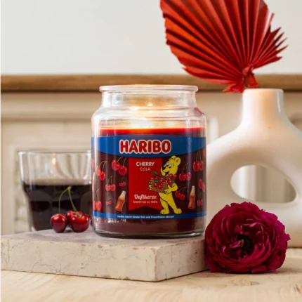 Haribo Cherry Cola Duftkerze brennt dekorativ neben Vase, Kirschen und roter Blüte