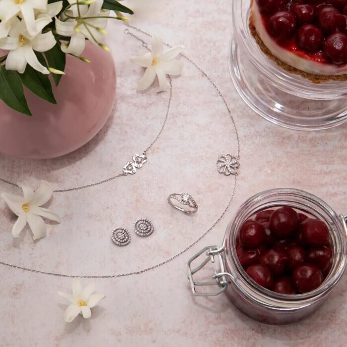 Schmuckstücke und Kette neben Kirschglas, Blumen und Kirschkuchen