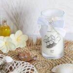 Fleur de Monoi Kerze mit brennendem Docht und Schmuckstück am weißen Schleifenband