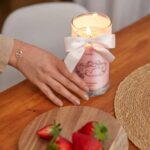 Brennende rosa Kerze mit Schleife neben Erdbeeren und einer Hand
