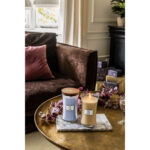 Brennende Gilded Sands Kerze neben einer violetten WoodWick Kerze auf Marmorsockel