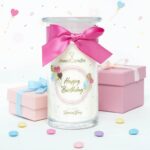 Happy-Birthday-Kerze mit rosa Schleife neben mehreren Schmuckketten mit Anhängern