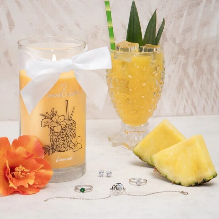 Jewel Candle L’Ananas neben Ananasgetränk, Ananasscheiben und orangefarbener Blüte
